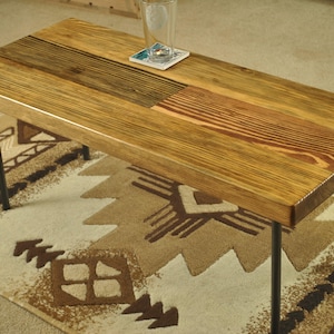 Peut inclure: Table basse rectangulaire en bois avec une finition naturelle, soutenue par des pieds en métal noir. Le plateau présente un mélange de grains et de couleurs de bois. Un verre et un dessous de verre sont posés dessus. La table est sur un tapis à motifs.