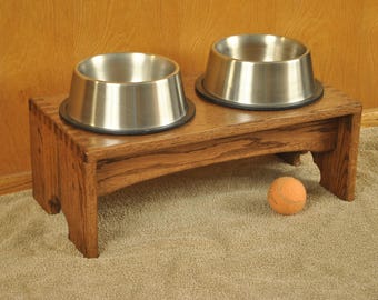 May include: A wooden pet feeder with two stainless steel bowls. The feeder is made of stained wood and has a small orange tennis ball resting on the floor in front of it. The bowls are elevated.