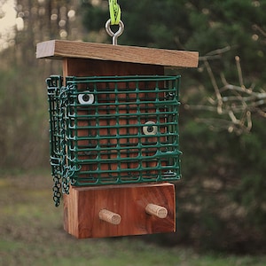 May include: A wooden bird feeder with a green wire cage and a wooden roof. The feeder has two wooden dowels and is suspended by a metal ring and a green strap. The background is a blurred view of trees and foliage.
