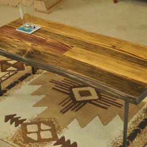 May include: A rectangular wooden coffee table with a natural edge and a warm, multi-toned finish. The table rests on four dark metal legs. A glass and coaster sit on the table, which is placed on a patterned rug.