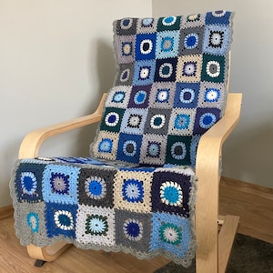 May include: A handmade crochet blanket and cushion cover set in shades of blue, gray, and white. The square pattern features floral-like designs. The blanket is draped over a light wood rocking chair, adding a cozy touch to the space.