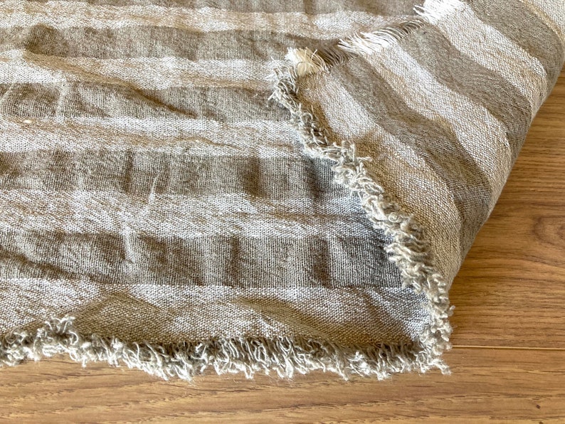 Puede incluir: Primer plano de una tela de lino a rayas marrones y blancas con un borde deshilachado. La tela est&aacute; doblada y tiene un aspecto suave y texturizado.