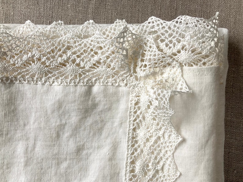 May include: A white linen tablecloth with a delicate white crocheted lace trim. The lace trim is intricate and has a scalloped edge.