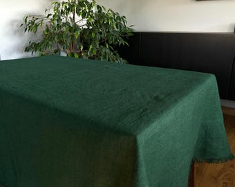 May include: A dark green rectangular tablecloth with a fringed edge. The fabric has a textured appearance and covers a wooden table. A leafy green plant is visible in the background.