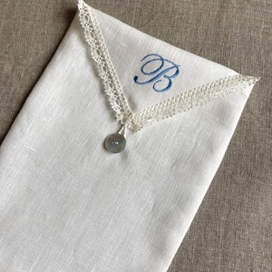 Puede incluir: Una bolsa de lino blanco con un borde de encaje color crema en forma de V en la parte superior. El centro de la bolsa está bordado con una letra B en azul. Un botón redondo de color gris translúcido está sujeto en la punta del encaje.