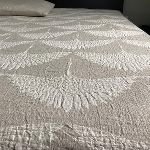 Può includere: Un copriletto beige con un motivo ripetuto di gru bianche. La trapunta è su un letto con un cuscino abbinato. Il motivo della gru è dettagliato e strutturato, creando un contrasto visivo. Il copriletto sembra morbido e invitante.