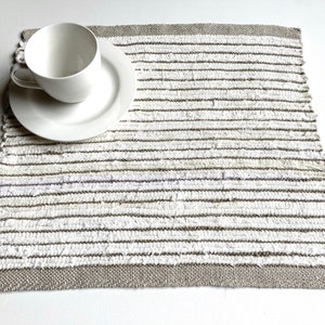 Op de afbeelding: Een witte kop en schotel liggen op een handgeweven placemat. De placemat heeft horizontale strepen in wit, grijs en bruin, met een grijze rand. De items zijn geplaatst op een wit oppervlak, wat een eenvoudige, neutrale uitstraling creëert.