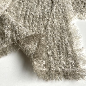 以下が含まれることがあります： フリンジのある薄い茶色のテクスチャのある生地のクローズアップ。生地は織り模様で、柔らかくふわふわした質感です。