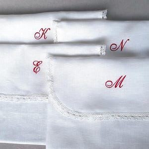 May include: White linen pouches with scalloped lace trim and embroidered red initials. The pouches are in various sizes and feature the letters K, E, N, and M. These pouches are ideal for storing small items.