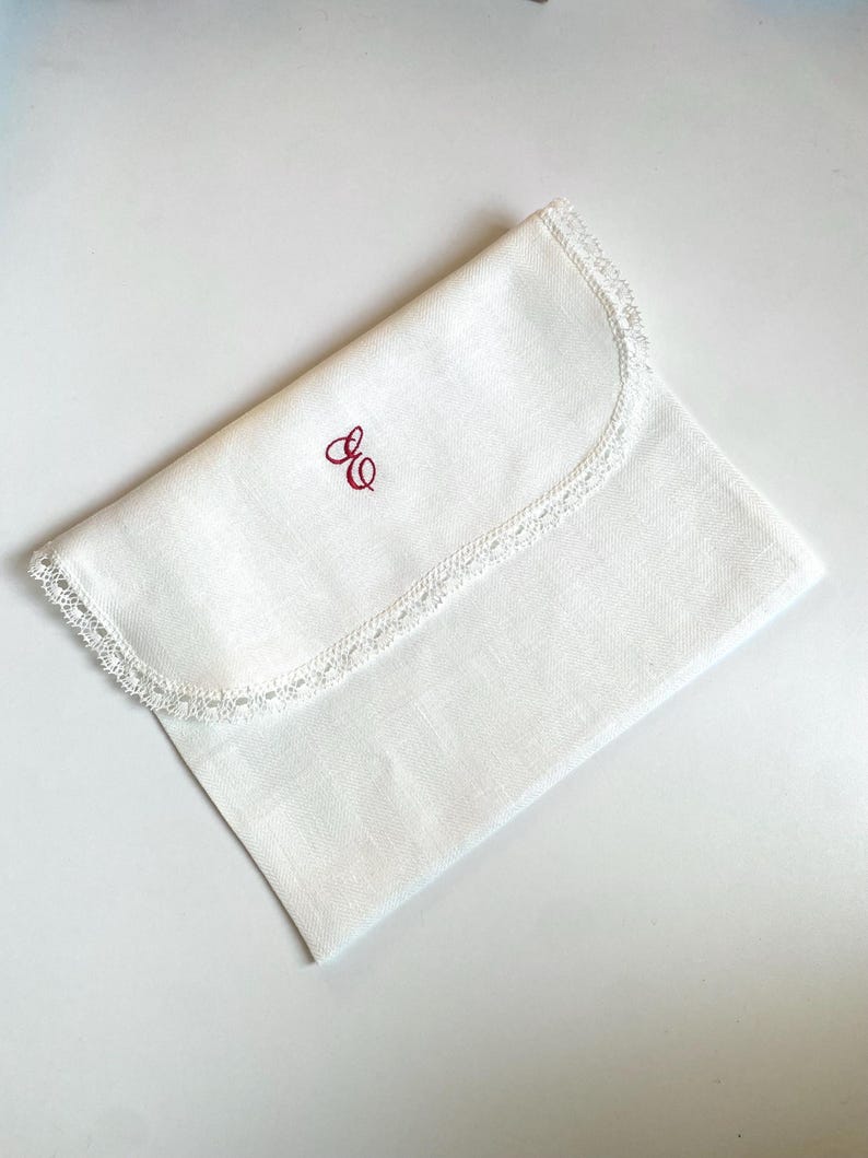 May include: A folded white linen pouch with a delicate lace trim along the curved edge. The pouch features a red embroidered monogram in the upper left corner. The item is displayed on a white surface.