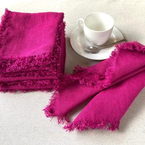 Może przedstawiać: Stos fuksjowych lnianych serwetek z postrzępionymi krawędziami. Na zdjęciu znajduje się również biała filiżanka i spodek z łyżeczką. Serwetki są złożone i wydają się nowe.