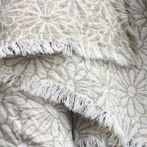 May include: A beige linen throw blanket with a white floral pattern and frayed edges.