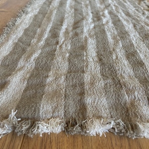Puede incluir: Un camino de mesa de lino beige con borde de flecos. El camino de mesa tiene un sutil patr&oacute;n de rayas.