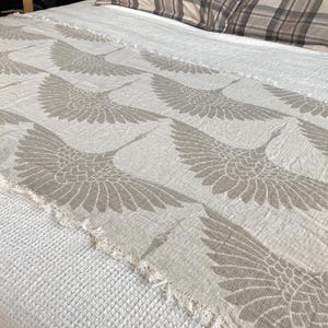 Pode incluir: Um corredor de cama de cor clara com um padrão repetido de guindastes estilizados em voo. Os guindastes são de uma cor cinza suave, contrastando com o fundo branco sujo. As bordas têm um detalhe com franjas.