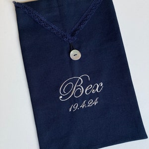 May include: Navy blue fabric pouch with a decorative lace trim and a mother-of-pearl button closure. The pouch is embroidered with the name "Bex" and the date "19.4.24" in a delicate script. The pouch is rectangular in shape.