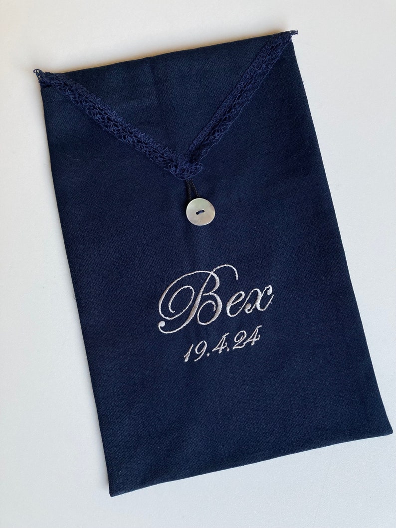 May include: Navy blue fabric pouch with a decorative lace trim and a mother-of-pearl button closure. The pouch is embroidered with the name "Bex" and the date "19.4.24" in a delicate script. The pouch is rectangular in shape.