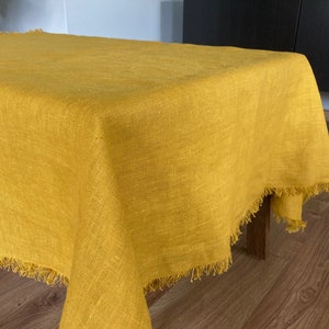 Könnte beinhalten: Eine gelbe Tischdecke aus Leinen mit Fransen. Die Tischdecke ist über einen Tisch mit einem Holzbein gelegt.