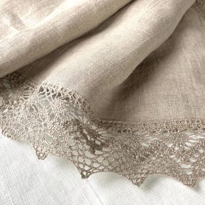 May include: A beige linen tablecloth with a delicate lace trim. The lace is a light brown colour and has a delicate floral pattern.