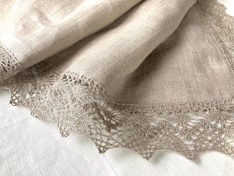 May include: A beige linen tablecloth with a delicate lace trim. The lace is a light brown colour and has a delicate floral pattern.