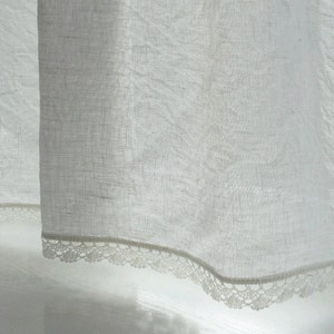 May include: White sheer curtain with a delicate lace trim. The curtain is hanging in a window and the light is shining through it.