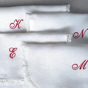 May include: White linen napkins with red embroidered initials: K, E, N, and M. Each napkin features delicate lace trim along one edge. The napkins are neatly folded, showcasing the elegant monogram details.