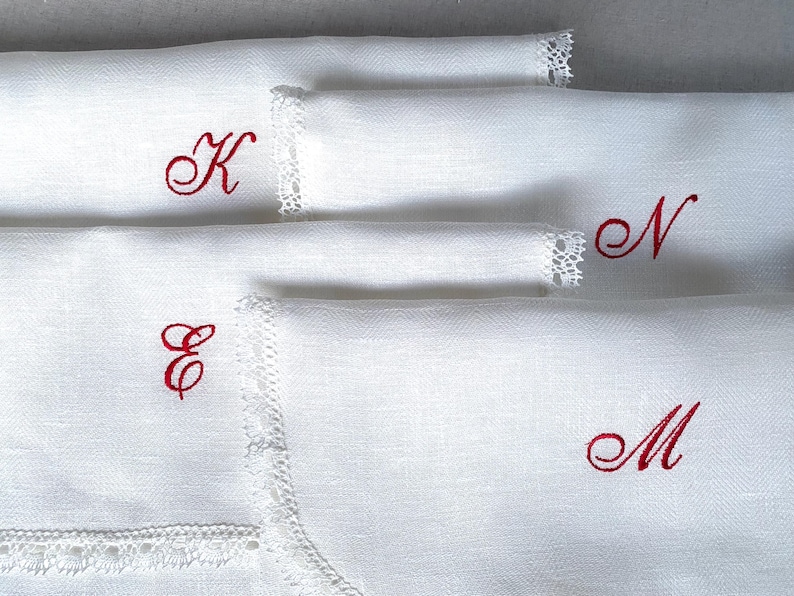 May include: White linen napkins with red embroidered initials: K, E, N, and M. Each napkin features delicate lace trim along one edge. The napkins are neatly folded, showcasing the elegant monogram details.