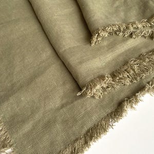 May include: Close-up of sage green linen fabric with a fringed edge. The fabric is folded, showing the texture and weave of the material. The fringed edges add a decorative element to the textile.