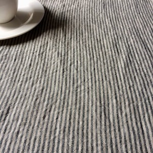 May include: A close-up of a striped linen tablecloth in shades of beige and black. The fabric has a subtle texture and a slightly wrinkled appearance.
