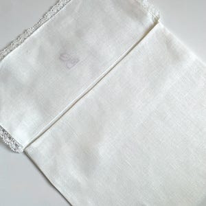 May include: A folded white linen napkin with a delicate lace trim along one edge. The napkin has a subtle herringbone weave pattern. The corner of the napkin has a faint, faded monogram. The overall aesthetic is simple and elegant.