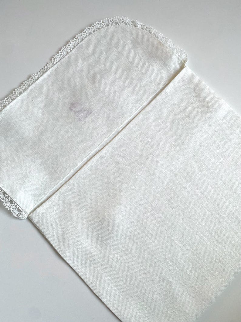 May include: A folded white linen napkin with a delicate lace trim along one edge. The napkin has a subtle herringbone weave pattern. The corner of the napkin has a faint, faded monogram. The overall aesthetic is simple and elegant.