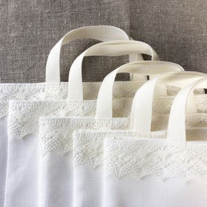 May include: Four white linen tote bags with lace trim. The bags have white canvas handles.