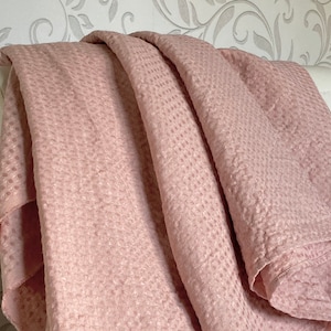 Puede incluir: Una manta suave de color rosa con un patrón de tejido de gofre texturizado. La manta está doblada y drapeada sobre una silla blanca.