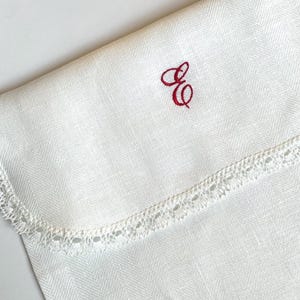 May include: Ivory-colored linen cloth with a herringbone weave pattern. The cloth features a delicate, scalloped lace trim along one edge. A red embroidered monogram of the letters "E" and "G" is in the upper right corner. The cloth is folded.