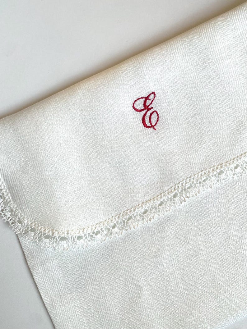 May include: Ivory-colored linen cloth with a herringbone weave pattern. The cloth features a delicate, scalloped lace trim along one edge. A red embroidered monogram of the letters "E" and "G" is in the upper right corner. The cloth is folded.