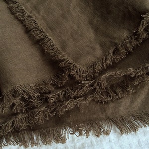 May include: A brown linen throw blanket with frayed edges. The blanket is folded in thirds and the edges are visible.