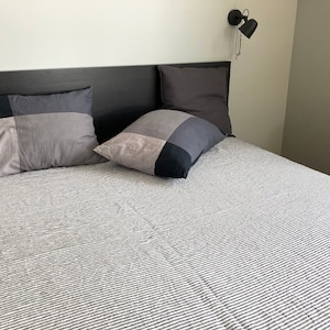 May include: A black bed frame with a white and grey striped bedspread and three grey pillows. The bed is against a white wall with a black lamp on the wall.
