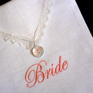 May include: White linen handkerchief with a delicate lace trim and a mother-of-pearl button. The word "Bride" is embroidered in a coral color. A classic wedding accessory.