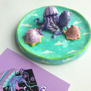 May include: A teal ceramic dish with a white rim and a yellow speckled glaze. The dish is decorated with white clouds and stars. Three small ceramic figurines of jellyfish and starfish are on the dish. The jellyfish are purple and pink, and the starfish are pink and yellow.