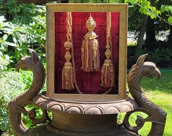 May include: A gold framed picture with a red velvet background and three gold tassels hanging from the top. The picture is sitting on top of a large, ornate, cast iron planter with two decorative handles shaped like dragon heads.