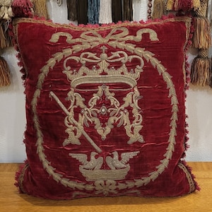 Puede incluir: Una almohada de terciopelo rojo con un diseño bordado en oro que presenta una corona, una cruz y un par de pájaros. La almohada tiene un borde con flecos.