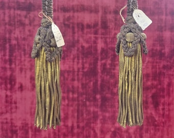 May include: Two gold and brown tassel tiebacks with a price tag attached to each. The tiebacks are hanging against a red velvet background.