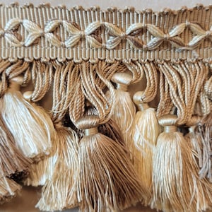 Puede incluir: Un ribete de flecos beige y crema con un diseño tejido. El ribete tiene una franja de flecos con una banda tejida en la parte superior.