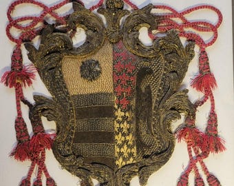May include: A gold and black embroidered coat of arms with red tassels and a red crown. The coat of arms is divided into four sections, each with a different design.