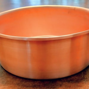 May include: A copper-colored bowl with a rounded shape and a smooth, reflective surface. The bowl has a slightly flared rim and appears to be made of metal. The interior is the same color as the exterior. The bowl is likely used for serving or display.