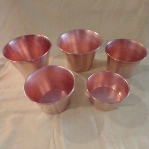 May include: A set of five copper-colored bowls of varying sizes arranged in a group. Each bowl features a smooth, metallic finish with a wide, open top and a tapered base. The bowls are circular in shape and have a consistent, polished appearance.