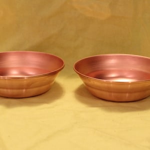 May include: Two copper bowls with a shiny, reflective surface. The bowls have a rounded shape with a slightly flared rim and a series of concentric rings around the exterior. The bowls are displayed against a yellow background.