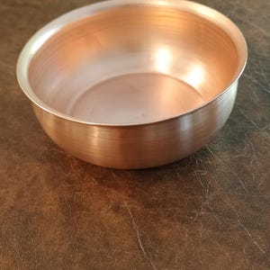 May include: A small, copper-colored bowl with a rounded shape. The bowl has a smooth, polished interior and a slightly textured exterior. The bowl is sitting on a brown surface.
