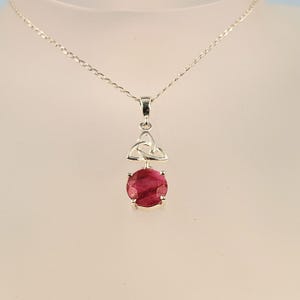 May include: A silver necklace featuring a round, faceted deep pink gemstone pendant suspended from a silver chain. Above the gemstone, a silver triquetra knot design connects the stone to the chain, creating a cohesive metallic and stone jewellery piece.