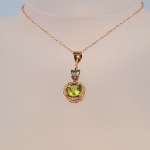 以下が含まれることがあります： 緑色の宝石のペンダントが、小さな透明な宝石で囲まれている、金のネックレス。ペンダントは繊細な金のチェーンに繋がっています。