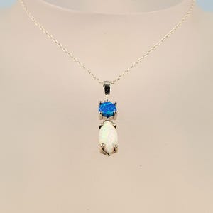 Op de afbeelding: Een zilveren ketting met een hanger met een blauwe edelsteen en een witte opaal. De blauwe edelsteen is rond en de opaal is druppelvormig. De hanger hangt aan een fijne zilveren ketting.